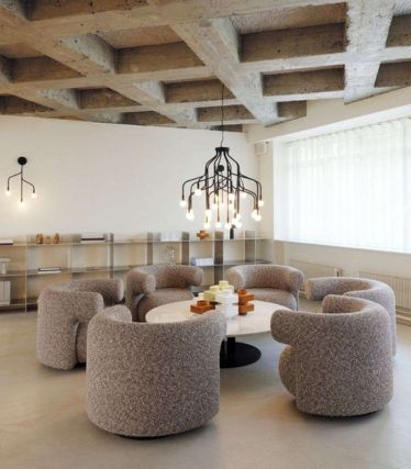 A modern interior with the Veskt pendant lamp by Normann Copenhagen as the central eye-catcher. The sculptural black lamp with several curved arms and freely hanging light bulbs illuminates a round table surrounded by six upholstered lounge chairs. Raw concrete ceilings, clean lines, and soft colors create a harmonious, contemporary ambience.
