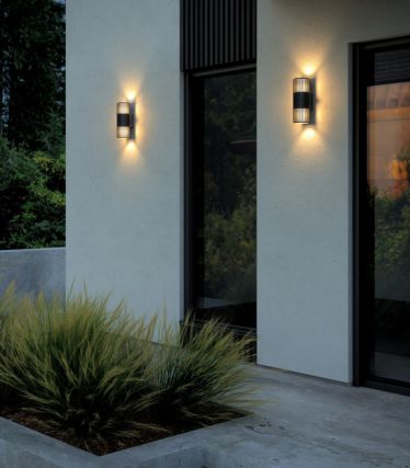 Modern exterior wall with two Aludra Double wall lights from Nordlux. The lights shine their light upwards and downwards, creating an elegant, warm play of light on the bright façade. In front of the house, green ornamental grasses and clean lines create a stylish, architectural ambience.