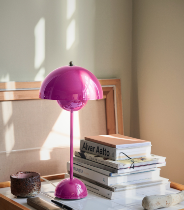 A bright pink table lamp with a glossy dome-shaped shade stands on a desk beside a stack of design books, including titles by Alvar Aalto. Soft daylight falls across the scene, highlighting the lamp and casting gentle shadows on the neutral background.
