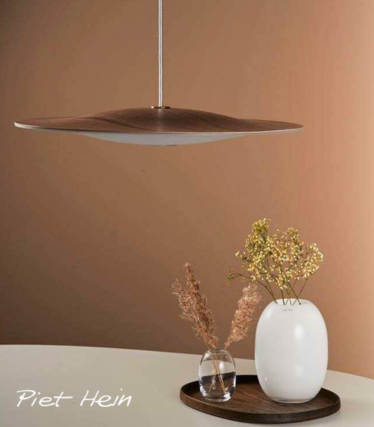 A minimalist pendant lamp with a thin, sculptural wooden shade hangs above a round table. On the table sits a wooden tray holding a white ceramic vase with delicate yellow flowers and a small glass vase with dried grasses. The scene is set against a warm beige background, creating a calm, natural atmosphere.