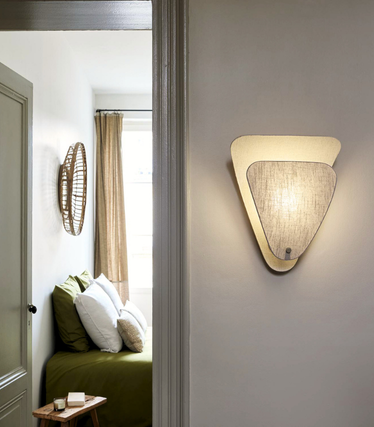 View through an open door into a quiet bedroom with a green bed. A triangular wall light made of natural materials with a textile lampshade lights up the wall.