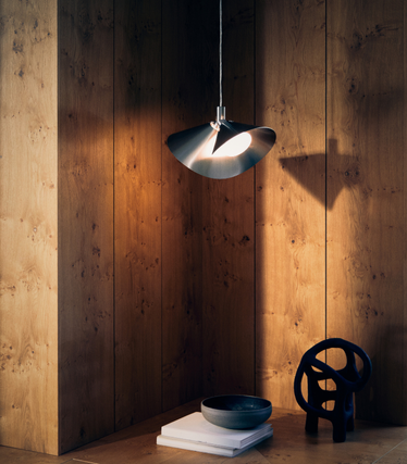Frandsen's brushed metal pendant light shines warmly in front of wood panelling. Beneath it are a stack of books, a bowl and a dark design sculpture on a wooden surface.