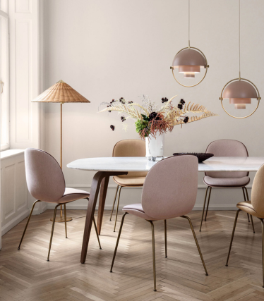 A soft, elegant dining room with a white marble table and pastel pink and beige chairs on brass legs. Over the table hang two round pendant lamps in warm beige tones with brass details. A tall floor lamp with a woven shade stands by the window, and a vase with dried floral arrangements decorates the table. The room features light herringbone wooden flooring and a calm, neutral colour palette.