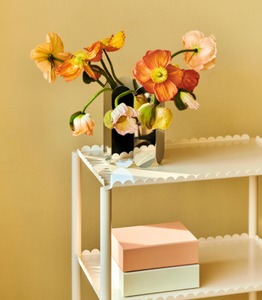 Orange and pale pink flowers in a reflective vase by HAY on a white trolley against a warm, soft background.