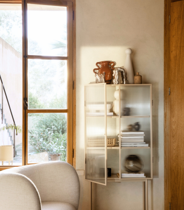 Bright living room corner with a display cabinet from Ferm Living, decorated with ceramic objects and books in warm daylight.