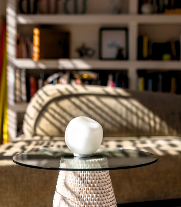 Small white table lamp on a glass top, with sunlight and a bookshelf in the background.