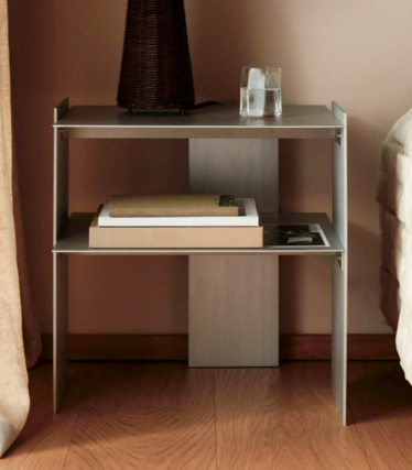 Slim metal bedside table with two shelves, books, a glass and a vase beside a bed.
