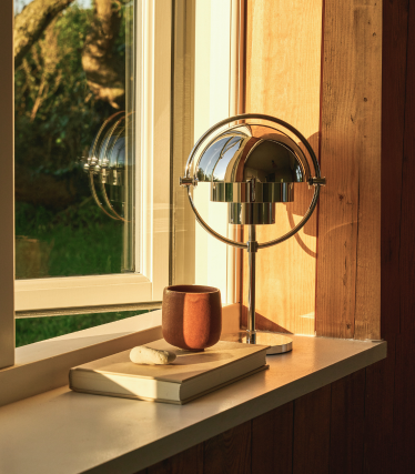Chrome table lamp on a windowsill in the evening light, next to a book and a ceramic mug.