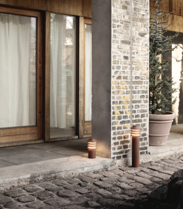 Outdoor scene with two modern cylindrical path lights placed beside a stone walkway and a brick-and-concrete column. Behind them are wooden-framed glass doors with light curtains, and a large potted evergreen plant. The warm glow of the lights creates a cozy and inviting atmosphere.