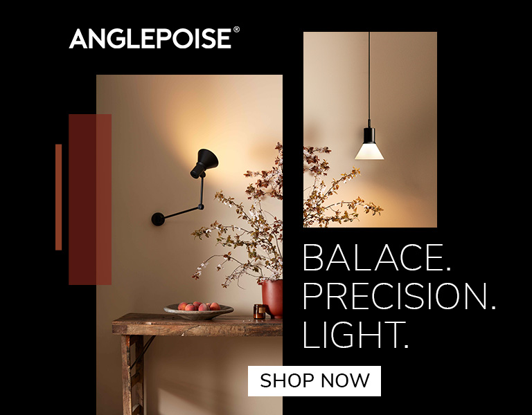 A black Anglepoise wall lamp illuminating a rustic wooden table with a bowl of fruit and a vase of branches. Another minimalist pendant light hangs on a beige wall. Text reads: ‘Anglepoise. Balance. Precision. Light.’ A button says ‘Shop Now.